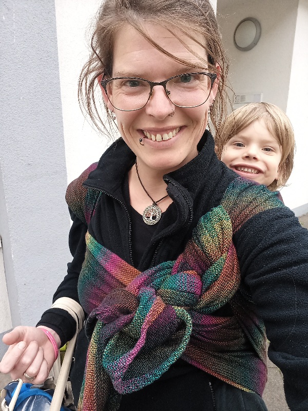 Ein Frau trägt ein 5 Jahre altes Kind in einem Regenbogen bunten Tragetuch auf dem Rücken. Beide lachen in die Kamera.