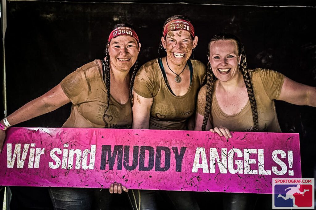Drei Frauen lachen und sind dreckig. Sie halten ein Schild mir der Aufschrift: "Wir sind Muddy Angel" 
