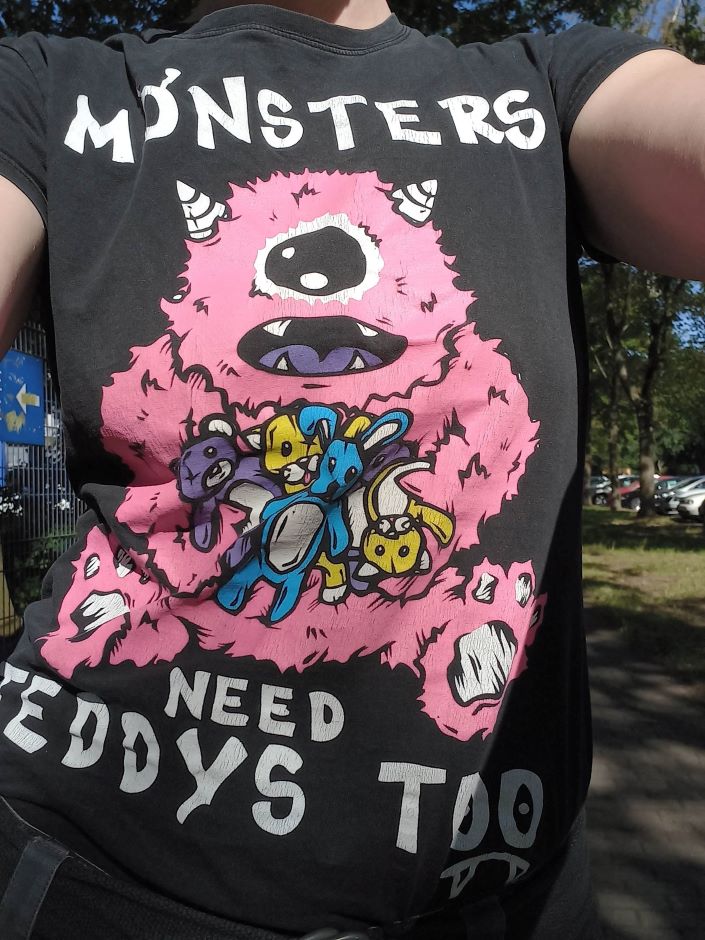 Das Bild zeigt ein Tshirt mit einem pinken Monster, das viele bunte Kuscheltiere hält. Der Schriftzug lautet "Monsters need Teddys, too"